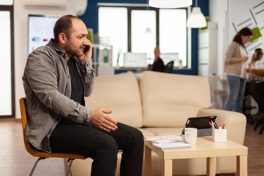 Nervous manager aggressively talking on phone and curses, screaming sitting on chair in middle of business office. Multiethnic scared coworkers planning financial project working in modern company