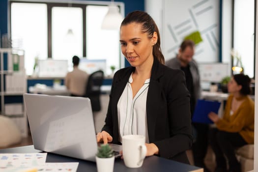 Enthusiastic woman manager typing on laptop, browsing on internet while sitting at desk smiling receiving good news. Multiethnic coworkers talking about startup financial company in modern office