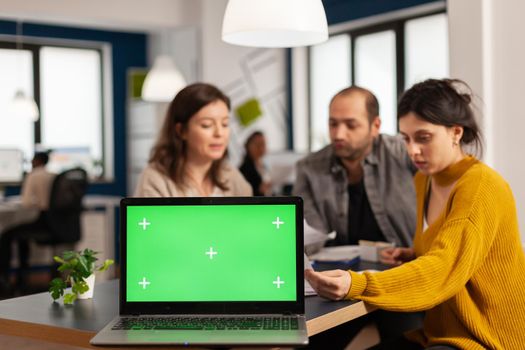 Laptop with green screen chroma key display. mock up desktop ready for presentation placed on desk while business people work in background. Group of employees using notebook with isolated monitor