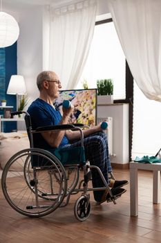 Invalid senior man in wheelchair holding workout dumbbells practicing arms exercise working at muscles body resistance training in living room. Pensioner looking at online workout course on tablet