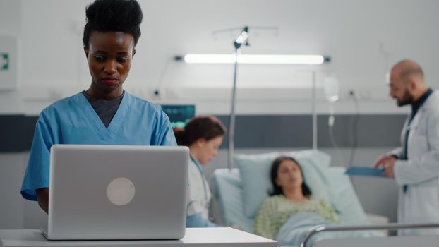 Afro american assistant typing sickness symptom on computer while specialist practitioner doctors monitoring sick woman writing recovery treatment on clipboard. Patient resting in bed in hospital ward