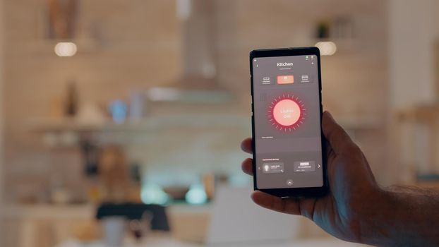 Close up of man hand holding phone with modern software controlling lights in house with automation lighting system, turning on bulb. Person looking at intelligent gadget sitting kitchen of smart home