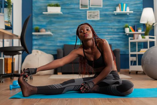 Fit black woman stretching reaching toes sitting on yoga mat after intense workout in home with dumbbells training. Relaxing muscles for healthy lifestyle wellbeing.