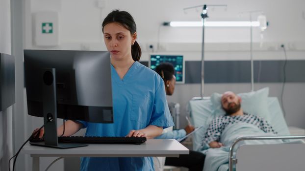 Front view of nurse typing medical expertise on computer while in background black practitioner doctor discussing with sick man healthcare treatment. Hospitalized patient having respiratory disorder