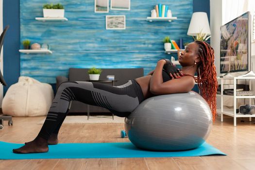 Fit sporty strong black woman working out core abdominals doing crunch exercise using swiss stability ball, on yoga mat in home living room dressed in sport wear and concentration on corect breathing.