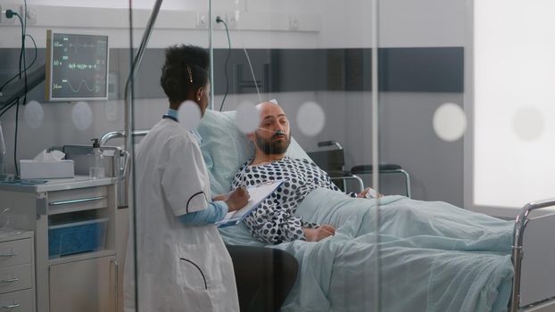 Practitioner specialist with black skin checking sick man writing disease treatment on clipboard monitoring disease symptom. Medical nurse arrange patient bed in hospital ward during recovery