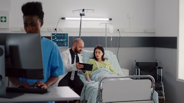 Practitioner doctor explaining disease diagnosis to sick woman using tablet computer. Patient resting in bed recovering after surgery in hospital ward while black nurse typing medical expertise