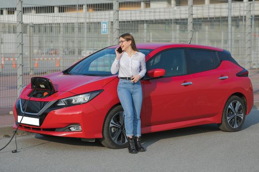 Girl stands with phone near her red electric car and waits when vehicle will charged. Plugging in power cord to an electric car. Ecological car connected and charging batteries.