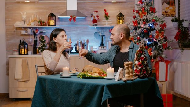 Man and woman clinking glasses of champagne and enjoying traditional meal at festive dinner on christmas eve. Couple celebrating seasonal holiday eating chicken and drinking alcohol