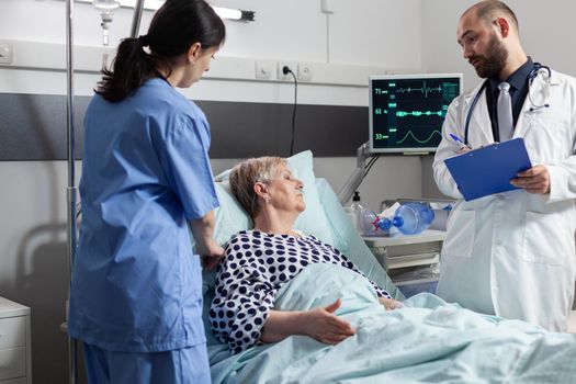 Senior woman laying in hospital bed receiving medication through an intravenous line, discussing with doctor about treatment and diagnosis. Using modern technology for therapy.