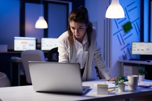Focused tired businesswoman reading emails on laptop standing near desk in start-up company late at night. Focused employee using technology network wireless doing finance research in office