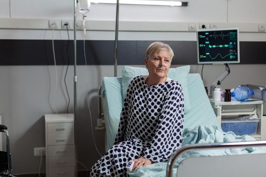 Portrait of sick senior patient getting recovery treatment from intravenous line sitting on hospital bed, breathing through oxygen mask because of pulmonary failure.