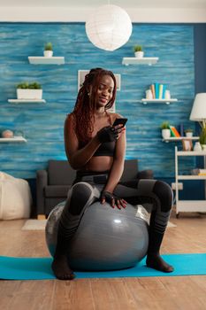 Active athletic black woman chatting on smartphone sitting on swiss ball in home living room, after working out on yoga mat to get stronger body and healthy lifestyle.