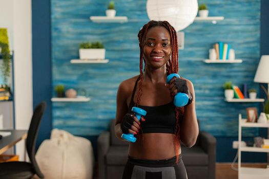 Athletic fit strong black woman working out with with dumbbells standing in home living room for healthy lifestyle, taking care of body exercising, using modern equipment.