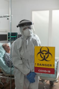 Nurse with face mask and coverall in hospital room, looking tired at camera, during coronavirus outbreak, to prevent infection. Patient laying in bed getting intravenous medicine treatment.