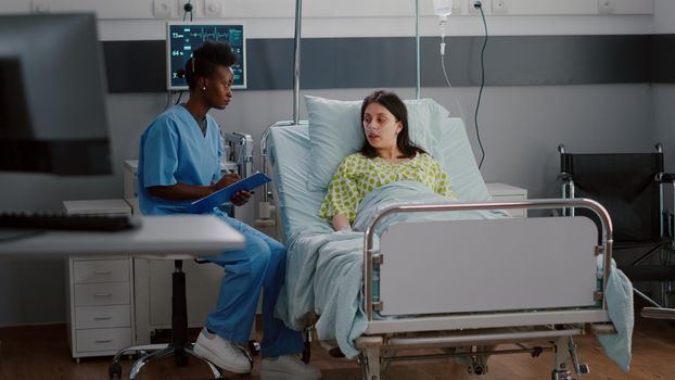 Afro-american nurse discussing with sick woman monitoring illness symptom writing healthcare treatment on clipboard. Sick woman resting in bed recovering after medical surgery in hospital ward