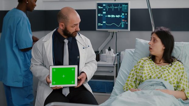 Specialist man in white coat talking with sick woman about illness recovery treatment. Practitioner holding mock up green screen chroma key tablet computer with isolated display in hospital ward