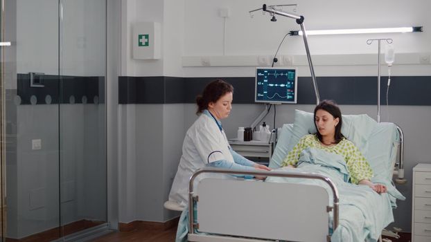 Specialist doctor putting oximeter checking pulse talking with sick woman explaining sickness symptom during recovery appointment. Patient resting in bed waiting for disease treatment in hospital ward