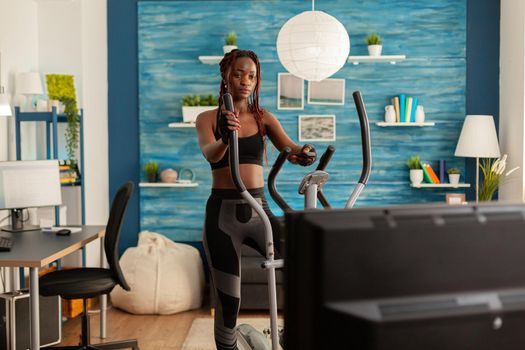 African fit strong woman doing cardio exercise on eliptical machine, in home living room looking at tv, watching instructions holding remote control. Exercising dressed in sportwear.