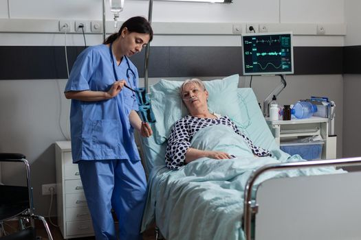 Medical nurse analyzing senior patient chest x-ray in hospital room, discussing explaining diagnosis. Old woman breathing through respiratory tube laying in bed listening health care specialist.