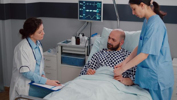 Sick man resting in bed wearing oxygen tube explaining disease symptom to practitioner doctor while writing sickness treatment on clipboard. Medical nurse putting oximeter examining recovery