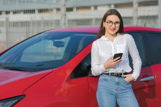 Girl stands with phone near her red electric car and waits when vehicle will charged. Plugging in power cord to an electric car. Ecological car connected and charging batteries