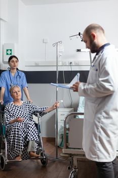 Senior woman getting medicine through intravenous line bag sitting on wheechair pushed by nurse in hospital room. Medical physician ward asking about pain to set diagnosis.