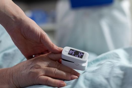 Close up of oxymeter attached on sick senior patient laying in hospital bed, monitoring hemoglobin. Medical staff using modern technology for reading blood oxygen saturation.