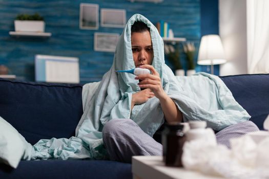 Young woman with flu symptoms checking temperature at home using thermometer. Adult measuring fever sitting on sofa in blanket. Tired unhealthy human in pain taking medical treatment