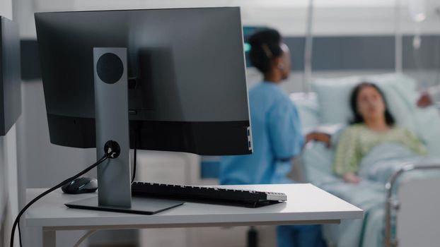 Hospitalized woman resting in bed while asisstant with black skin monitoring sickness symptom writing medical expertise on clipboard. Assistant discussing illness therapy working in hospital ward