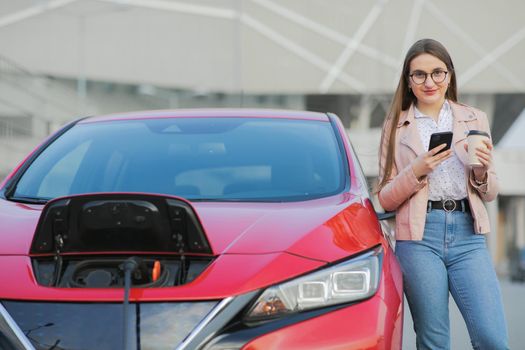 Girl stands with phone near her electric car and waits when vehicle will charged. Charging of electric car.