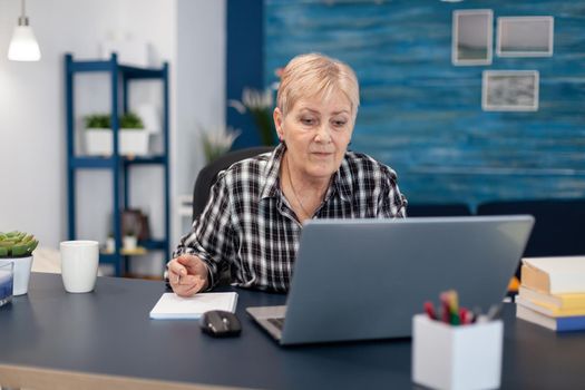 Senior entrepreneur reading from portable computer without glasses. Elderly woman in home living room using moder technoloy laptop for communication sitting at desk indoors.