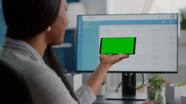 African american student working remote from home looking with mock up green screen chroma key phone with isolated display sitting at desk in living room. Black teenager browsing social network