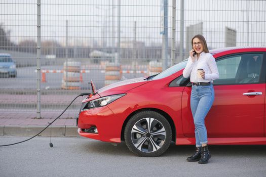 Ecological Car Connected and Charging Batteries. Girl Use Coffee Drink While Using SmartPhone and Waiting Power Supply Connect to Electric Vehicles for Charging the Battery in Car