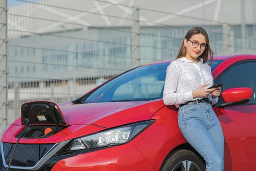 Caucasian girl using smart phone and waiting power supply connect to electric vehicles for charging the battery in car. Ecological car connected and charging batteries