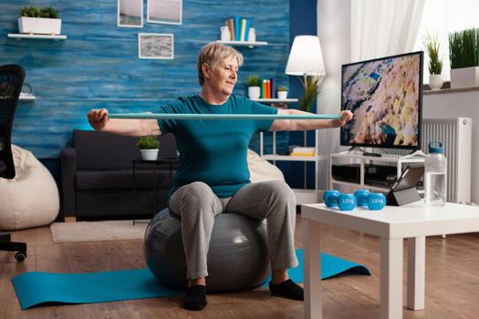 Retired senior man sitting on fitness swiss ball in living room doing wellness fitness workout streching arm muscles using aerobics elastic band. Pensioner training body resistance in living room