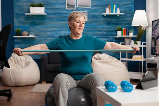 Retired cheerful pensioner sitting on swiss ball watching fitness video on tablet streching arm using elastic band during healthcare workout in living room. Senior woman working body resistance