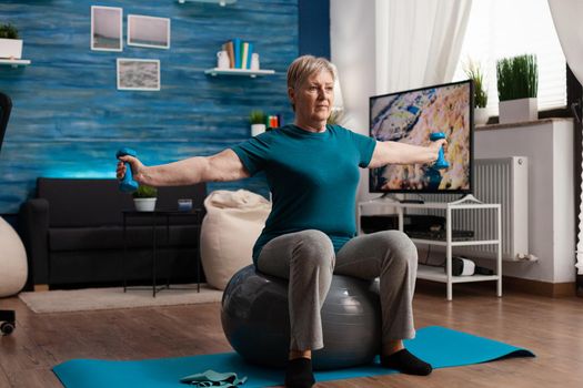 Focused senior woman stretching arm working at body muscle using fitness dumbbells sitting on swiss ball in living room. Caucasian male exercising muscular healthcare during wellness workout