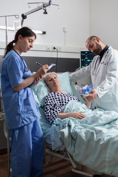 Doctor checking senior patient in hospital intensive care, checking oxygen saturation, blood pressure on oxymeter attached on sick senior patient while nurse making notes on clipboard for diagnosis.