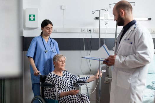 Elderly sick patient sitting in hospital wheelchair getting treatment through iv drip bag and oxymeter attached on finger monitoring oxygen saturation from blood. Doctor discussing about recovery therapy.