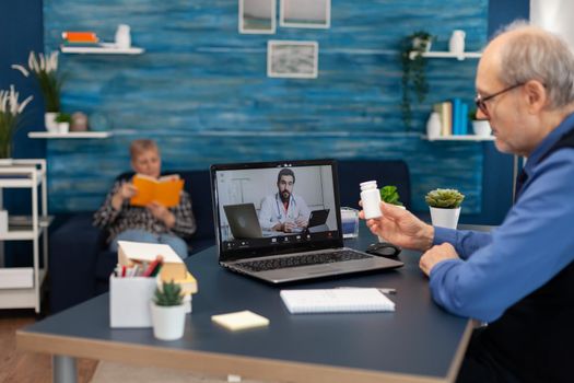 Young doctor examining senior man during video conference. Elderly man discussing with healthcare practitioner in the course of remote call and wife is reading a book on sofa.