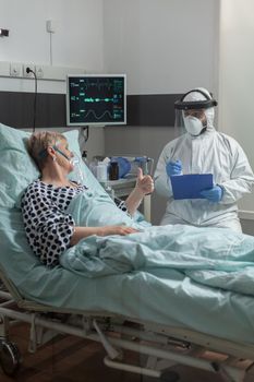 Doctor wearing ppe protection suit against contamination with covid-19, in hospital room during visit at senior woman laying in bed breathing with help from oxygen mask. and getting intravenous medicine.