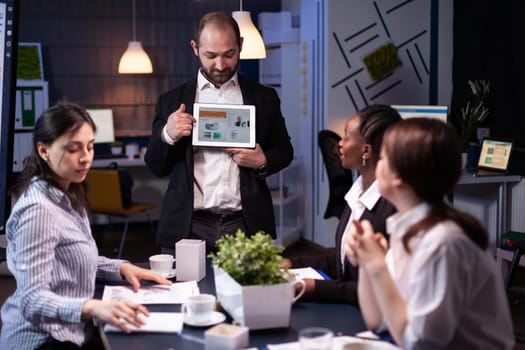 Focused businessman showing corporate graphs presentation using tablet working at company ideas. Diverse multi-ethnic businesspeople brainstorming strategy late at night in office meeting room