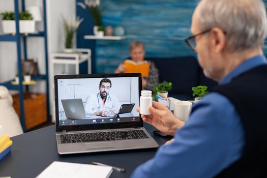 Senior man listening doctor during telemedicine holding pills bottle. Elderly man discussing with healthcare practitioner in the course of remote call and wife is reading a book on sofa.