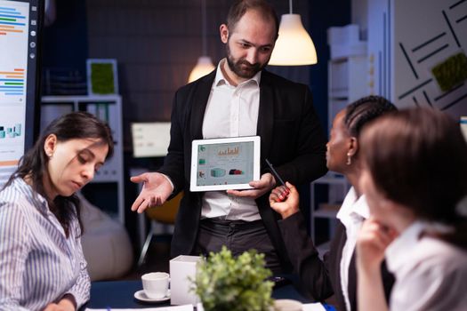 Focused workaholic entrepreneur man working overtime presenting company statistics using tablet. Diverse multi-ethnic businesspeople overworking in office meeting room late at night.