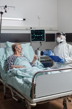 Doctor dressed in ppe suit with face shiled discussing with senior patient, laying in bed with oxygen mask during coronavirus outbreak. Getting intravenous medicine through iv drip bag.