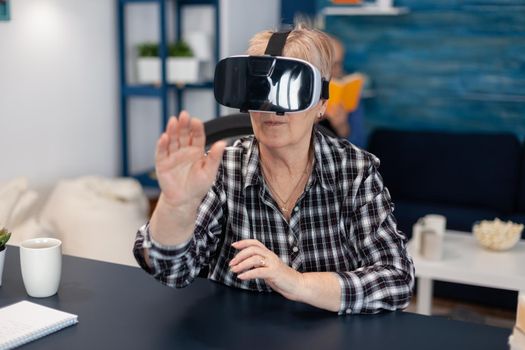 Happy senior woman wearing virtual reality headset doing hand gesture. Cheerful elderly woman experiencing augmented reality cyberspace while husband is reading a book on sofa.