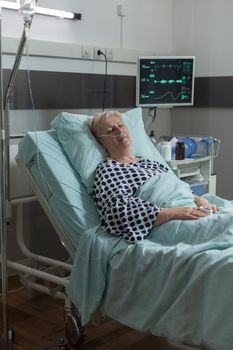 Senior woman resting in hospital bed breathing with help from oxygen mask, because of pulmonary illness. Getting intravenous medicine from iv drip bag during recovery therapy.