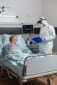Senior woman patient laying unconscious in hospital bed during coronavirus outbreak, breathing with help from oxygen mask. Doctor dressed in ppe suit to prevent infection.