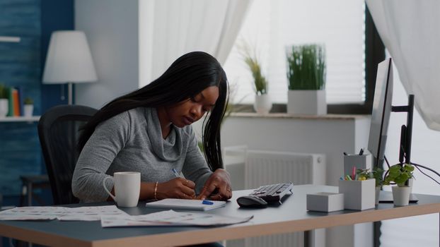 Black student writing education ideas on stickey notes sitting at desk table in living room. Young woman studying business at univeristy using elearning platoform working remote from home